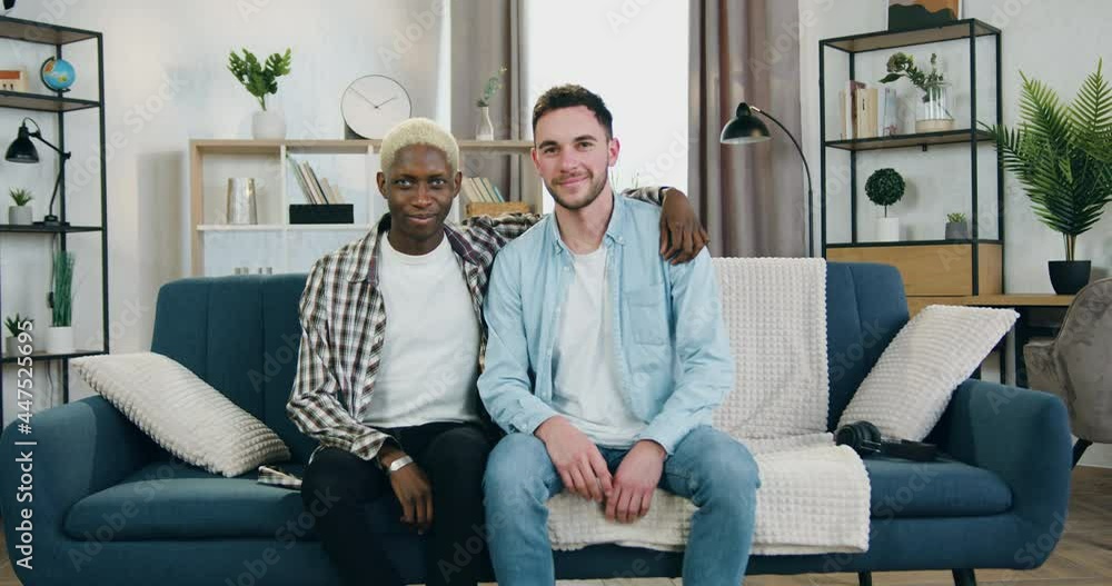 Likable happy young african american gay hugging his european boyfriend by the shoulder while they both sittting on the couch at home and looking at camera with smiles Likable happy young african american gay hugging his european boyfriend by the shoulder while they both sittting on the couch at home and looking at camera with smiles