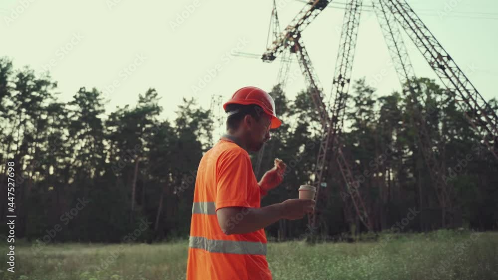 Hungry worker in overalls in electric power business eats sandwich and drinks takeaway coffee at workplace near power lines. Power worker eating snack with hot drink near high voltage power line. Hungry worker in overalls in electric power business eats sandwich and drinks takeaway coffee at workplace near power lines. Power worker eating snack with hot drink near high voltage power line.