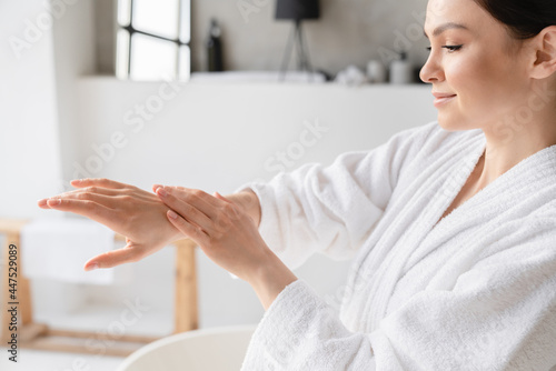 Fotografi Young caucasian woman in spa bathrobe applying hand cream moisturizer on her hands for softing effect