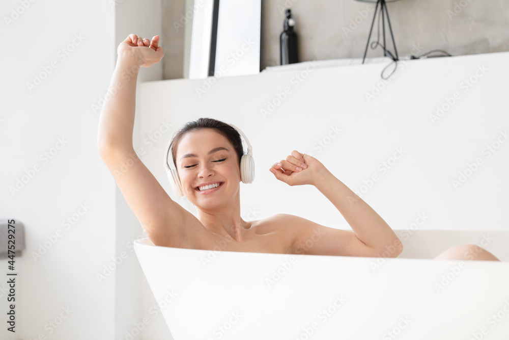 Smiling serene peaceful young woman girl taking bath shower at home listening to the music in headphones and dancing, relaxing in hot water after tedious day. Beauty treatment, body and skin spa Smiling serene peaceful young woman girl taking bath shower at home listening to the music in headphones and dancing, relaxing in hot water after tedious day. Beauty treatment, body and skin spa