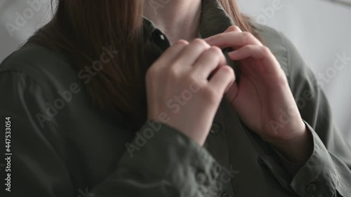 Young woman brunette with loose flowing hair unbuttons green-grey shirt with trembling hands standing at home close view.