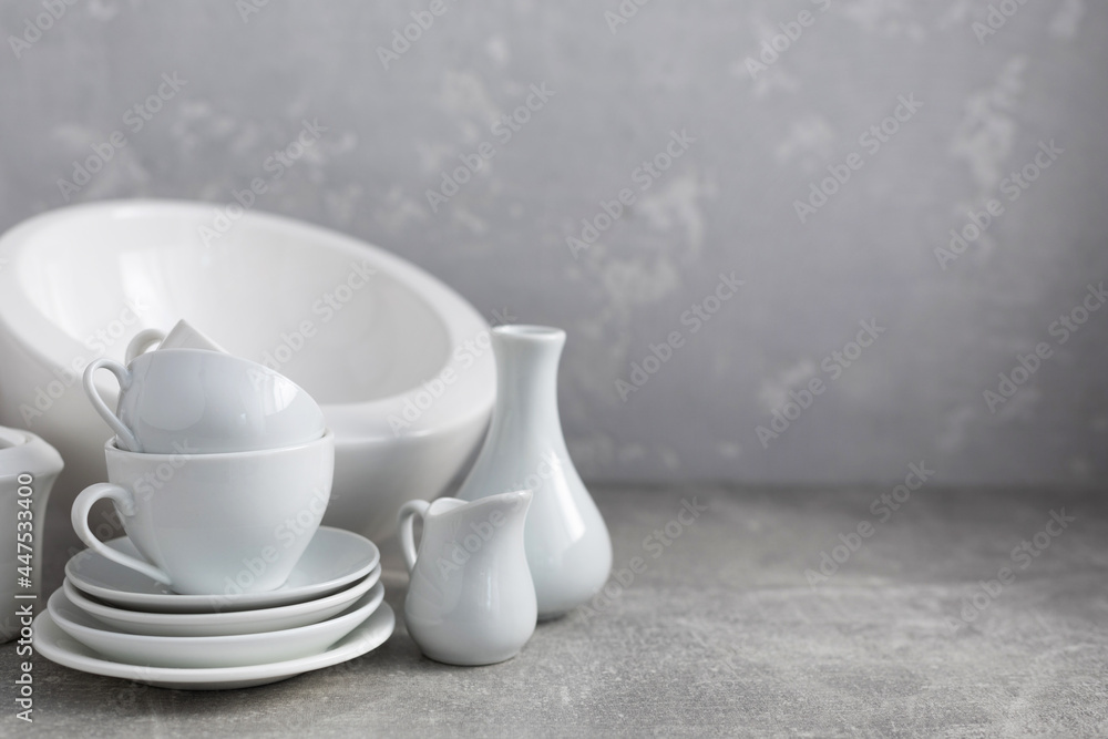 Empty crockery set or ceramic dishes. White kitchen dishware and tableware on table near grey wall Empty crockery set or ceramic dishes. White kitchen dishware and tableware on table near grey wall