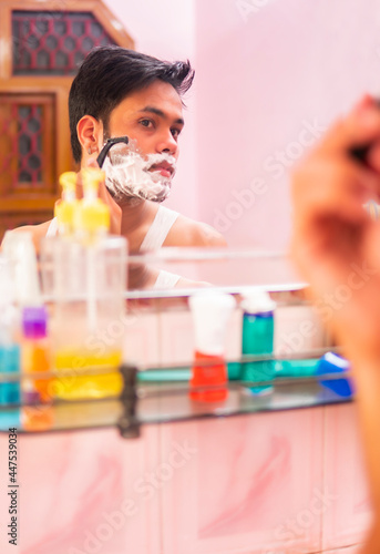Indian young man looking into mirror and shaving his facial hair with a razor.