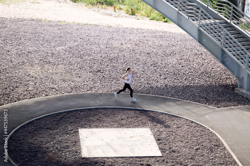 A young woman in a white T-shirt and black pants runs in a circle, illuminated by the evening sun. Running training in the city. Preparing for the competition.