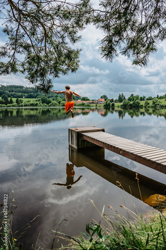 Rear view of man jumping from wooden pier with joy in to the water,nature reflected in lake.Having summer fun by pond.Travel vacation, youth holiday concept.Happy active lifestyle, entertaintment photo