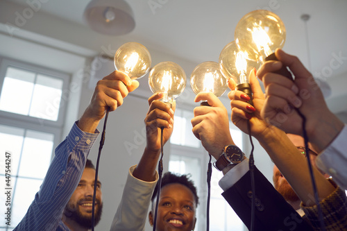 Happy smiling interracial group of people standing together with raised lightbulb office shot. Focus on illuminated glowing lamp. Creative thinking and teamwork, brainstorming and idea share concept photo