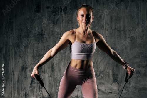 Middle shot portrait of muscular young athletic woman with perfect beautiful body in sportswear exercising with resistance band. Caucasian fitness female training with stretching expander in studio.