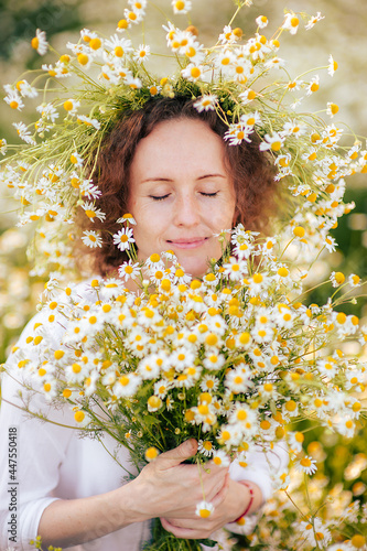Portrait of a happy woman with closed eyes in a white dress with a wreath of daisies on her head and a bouquet of flowers in her hands in a field of daisies