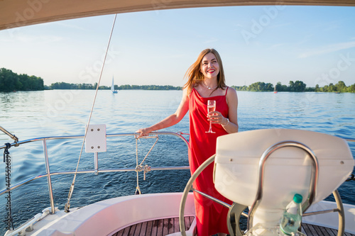 The elegant woman in a long red dress holding a glass of champagne standing on a yacht and looking for river sunset view © svittlana