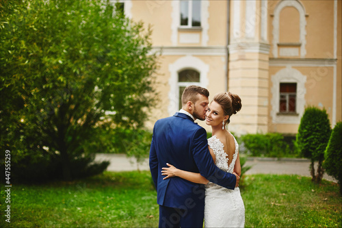 A handsome man in a trendy blue suit hugging and kissing a beautiful model woman in a wedding dress. A happy couple of newlyweds hugging after the outdoor wedding ceremony