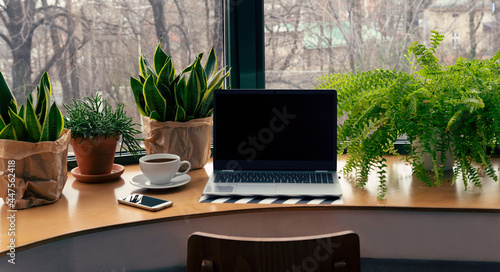 Office desk with green plants and laptop with mock up in vintage style. Workspace with window in modern interior. Technology, business with smartphone and computer in cafe. Banner.