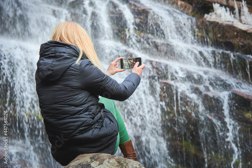 Young woman, a hiker or a camper, taking a picture of a big waterfall, sitting on a rock, view from behind; autumn weather, warm clothing
