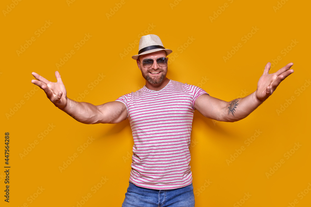 Come closer, let me hug you. Studio shot of positive friendly bearded man in trendy stripped outfit and hat pulling hands towards camera, inviting mates to come inside, welcoming guest to party