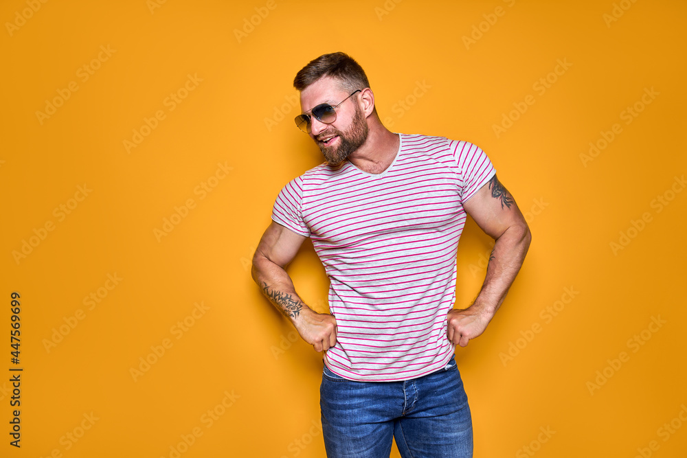 Muscular confident bearded male in shirt, jeans and stylish sunglasses standing in studio over yellow background Muscular confident bearded male in shirt, jeans and stylish sunglasses standing in studio over yellow background