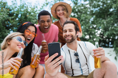 happy man taking selfie with blurred multiethnic friends during beer party
