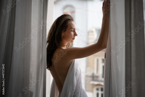Beautiful sensual young woman with long hair in a white shirt standing by the window with curtains in the apartment