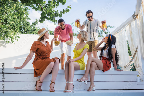 Quadro su tela Positive multiethnic men holding beer and food near friends in patio