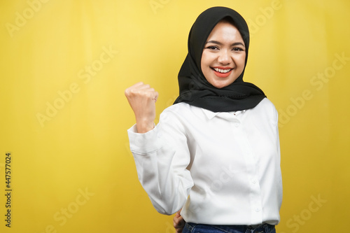 Beautiful young asian muslim woman smiling confident, enthusiastic and cheerful with hands clenched, sign of success, punching, fighting, not afraid, victory, isolated on yellow background © MunirSr