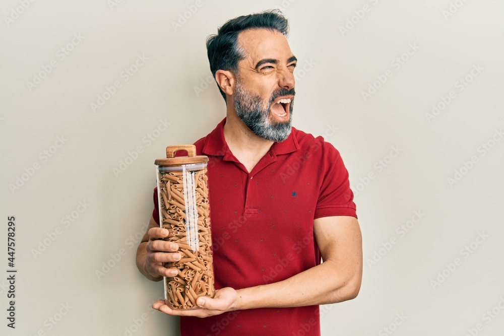 Fototapeta premium Middle age man with beard and grey hair holding jar with macaroni pasta angry and mad screaming frustrated and furious, shouting with anger. rage and aggressive concept.