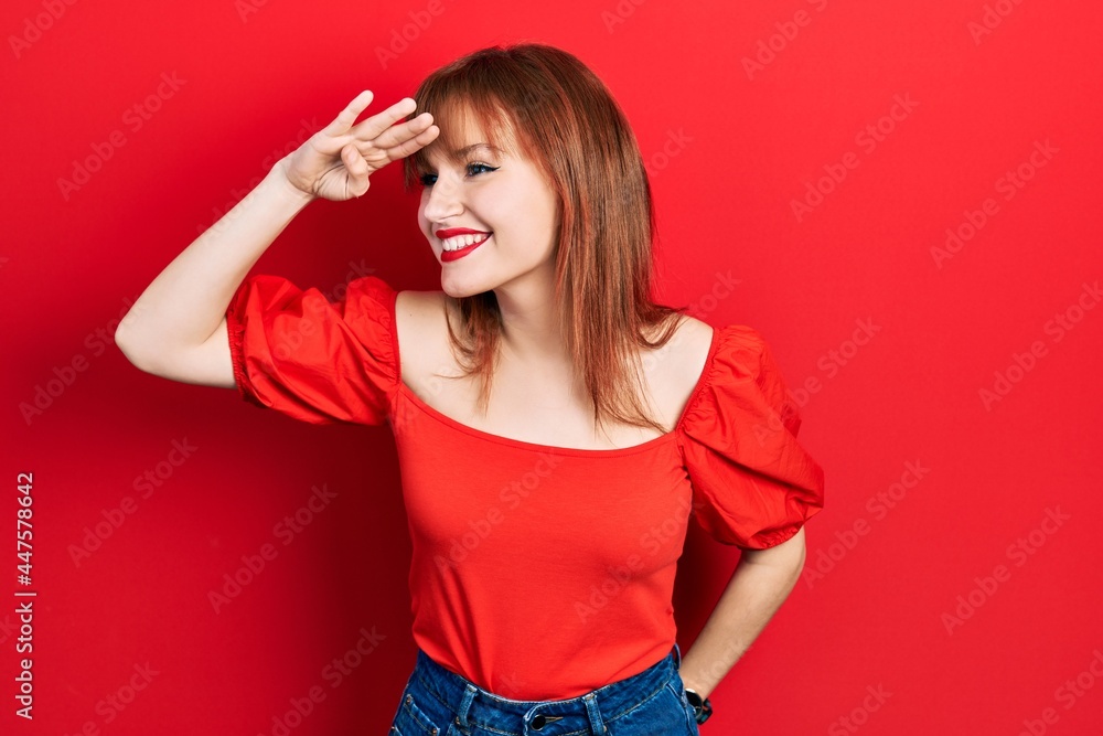Redhead young woman wearing casual red t shirt very happy and smiling looking far away with hand over head. searching concept. Redhead young woman wearing casual red t shirt very happy and smiling looking far away with hand over head. searching concept.
