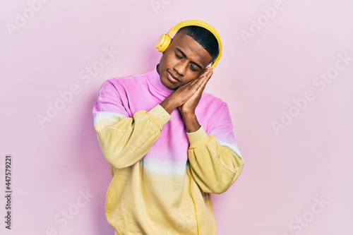 Young black man listening to music wearing headphones sleeping tired dreaming and posing with hands together while smiling with closed eyes.