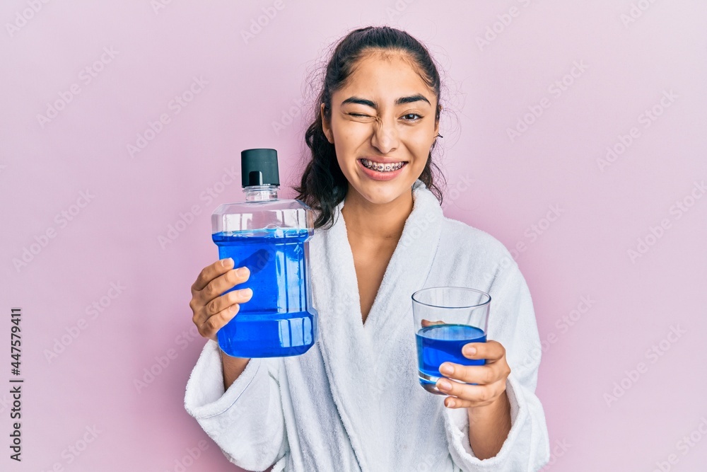 Hispanic teenager girl with dental braces holding mouthwash winking looking at the camera with sexy expression, cheerful and happy face. Hispanic teenager girl with dental braces holding mouthwash winking looking at the camera with sexy expression, cheerful and happy face.