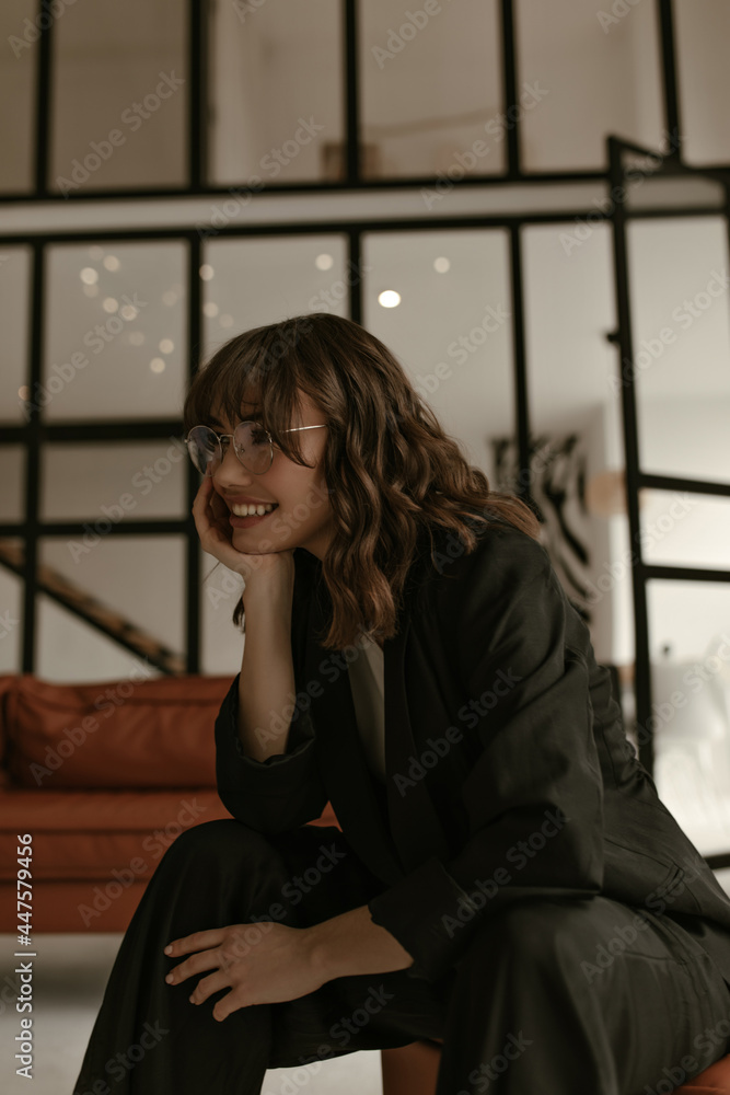 Happy brunette curly woman in black stylish jacket and silk dark pants smiles in living room. Lady in eyeglasses and suit sits on chair at home.
