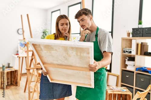 Young hispanic artist couple with serious expression holding canvas at art studio.
