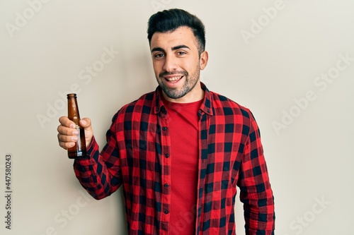 Young hispanic man drinking a bottle of beer looking positive and happy standing and smiling with a confident smile showing teeth