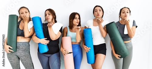Group of women holding yoga mat standing over isolated background looking at the camera blowing a kiss with hand on air being lovely and sexy. love expression.