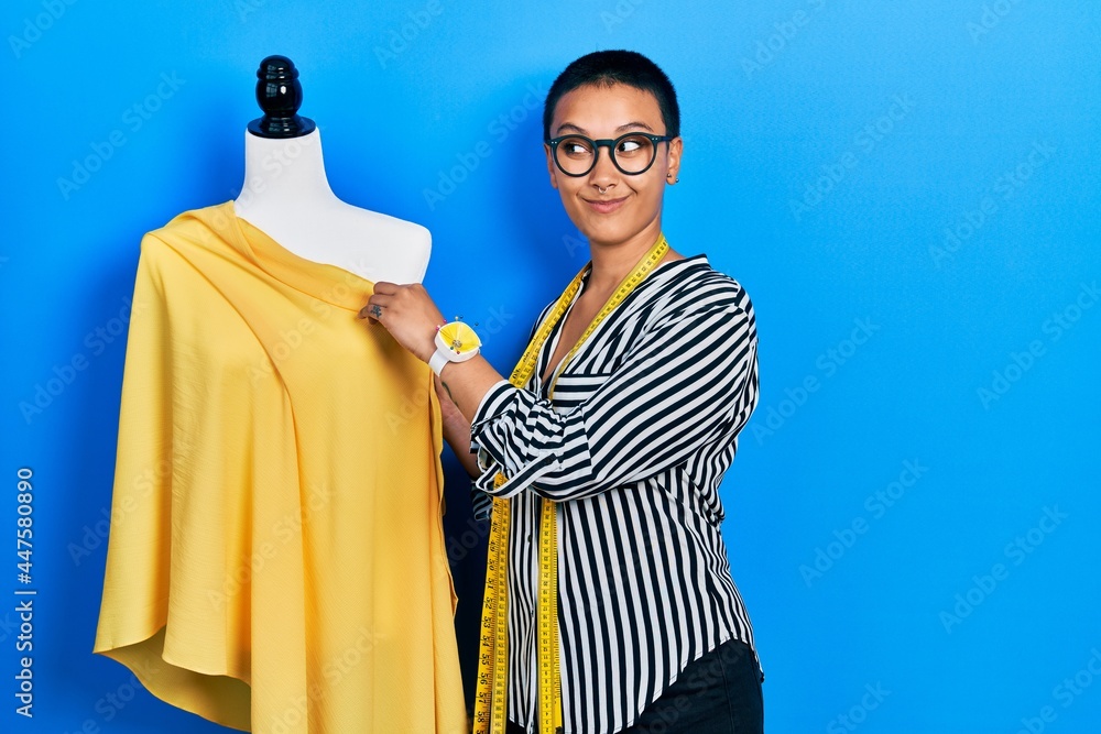 Fototapeta premium Beautiful hispanic woman with short hair standing by manikin sewing cloth smiling looking to the side and staring away thinking.