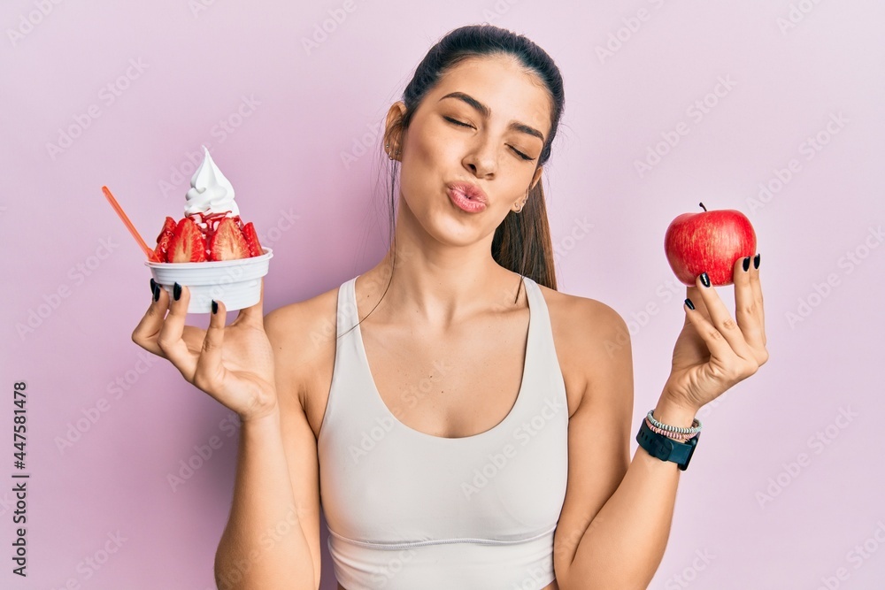 Young hispanic woman wearing sportswear holding apple and ice cream looking at the camera blowing a kiss being lovely and sexy. love expression. Young hispanic woman wearing sportswear holding apple and ice cream looking at the camera blowing a kiss being lovely and sexy. love expression.