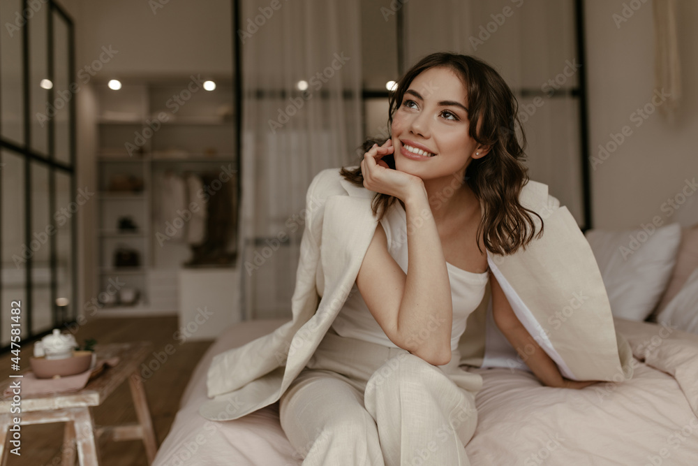 Tanned brunette woman in stylish pants and beige jacket sits on bed at home. Young charming lady in suit smiles in bedroom. Tanned brunette woman in stylish pants and beige jacket sits on bed at home. Young charming lady in suit smiles in bedroom.