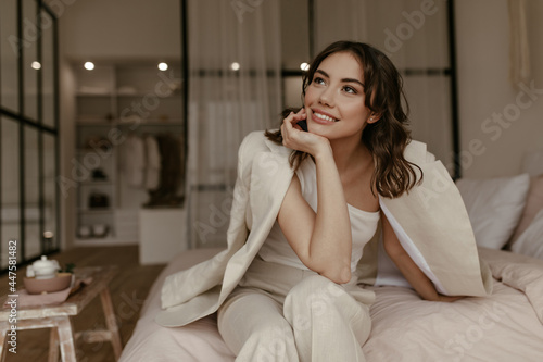 Fotografie Tanned brunette woman in stylish pants and beige jacket sits on bed at home