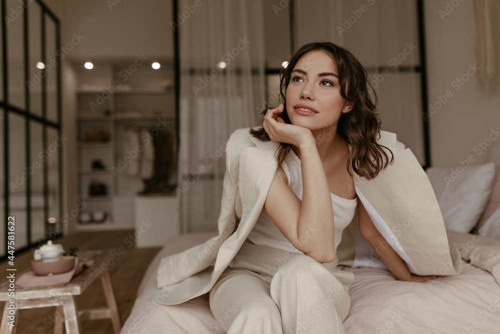 Curly brunette woman in beige pants and linen jacket leans on knee. Attractive girl in suit sits on soft bed in cozy light bedroom. Curly brunette woman in beige pants and linen jacket leans on knee. Attractive girl in suit sits on soft bed in cozy light bedroom.