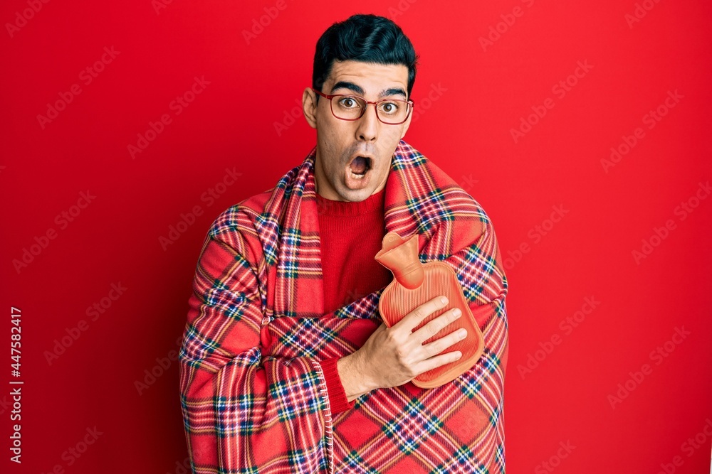 Handsome hispanic man wrapped in a winter blanket holding hot water bag afraid and shocked with surprise and amazed expression, fear and excited face. Handsome hispanic man wrapped in a winter blanket holding hot water bag afraid and shocked with surprise and amazed expression, fear and excited face.