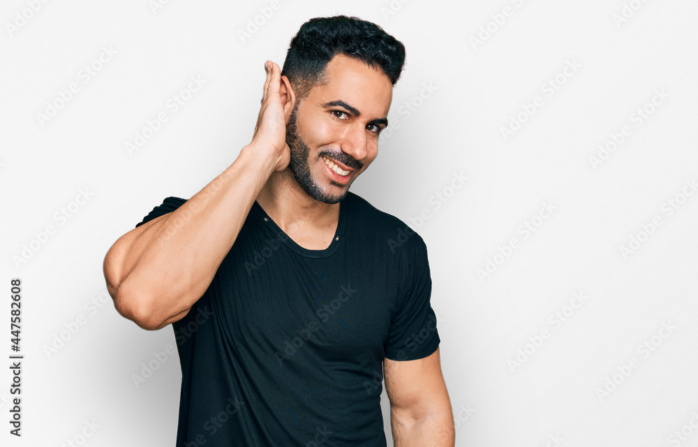 Hispanic man with beard wearing casual black t shirt smiling with hand over ear listening an hearing to rumor or gossip. deafness concept. Hispanic man with beard wearing casual black t shirt smiling with hand over ear listening an hearing to rumor or gossip. deafness concept.