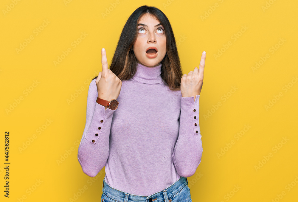 Young beautiful teen girl wearing turtleneck sweater amazed and surprised looking up and pointing with fingers and raised arms. Young beautiful teen girl wearing turtleneck sweater amazed and surprised looking up and pointing with fingers and raised arms.