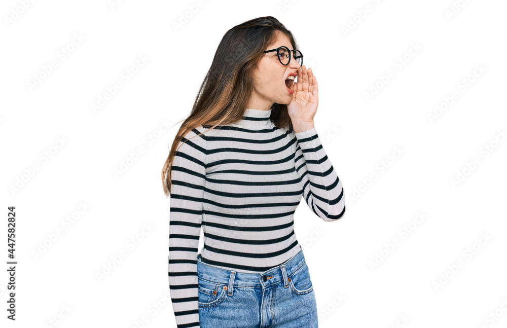 Young beautiful teen girl wearing casual clothes and glasses shouting and screaming loud to side with hand on mouth. communication concept. Young beautiful teen girl wearing casual clothes and glasses shouting and screaming loud to side with hand on mouth. communication concept.