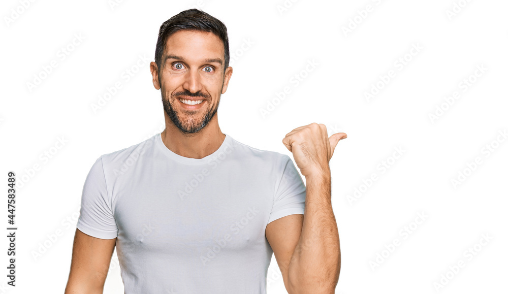 Handsome man with beard wearing casual white t shirt smiling with happy face looking and pointing to the side with thumb up. Handsome man with beard wearing casual white t shirt smiling with happy face looking and pointing to the side with thumb up.