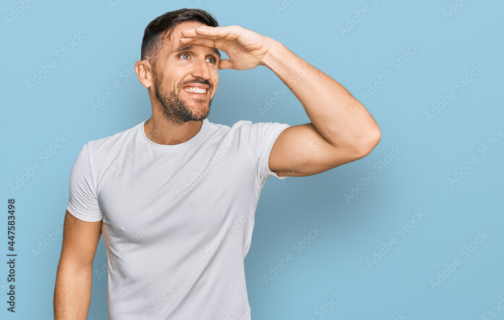 Handsome man with beard wearing casual white t shirt very happy and smiling looking far away with hand over head. searching concept. Handsome man with beard wearing casual white t shirt very happy and smiling looking far away with hand over head. searching concept.