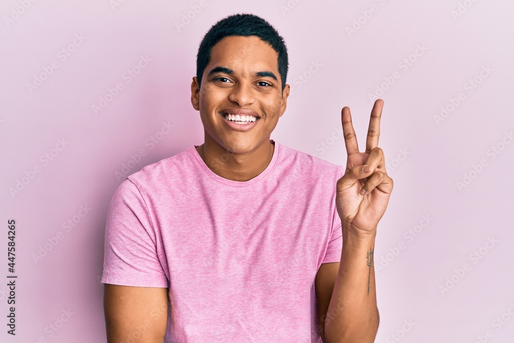 Young handsome hispanic man wearing casual pink t shirt smiling with happy face winking at the camera doing victory sign. number two. Young handsome hispanic man wearing casual pink t shirt smiling with happy face winking at the camera doing victory sign. number two.