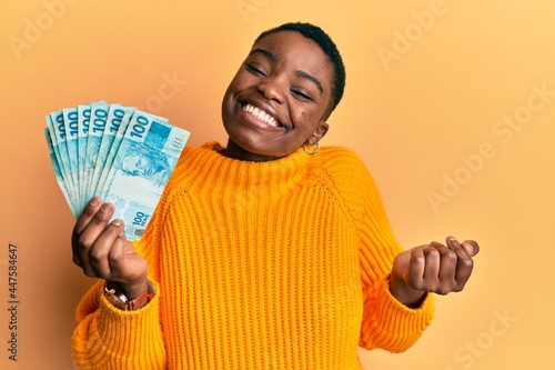 Young african american woman holding 100 brazilian real banknotes screaming proud, celebrating victory and success very excited with raised arm