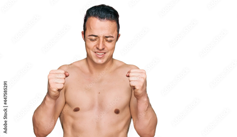 Handsome young man wearing swimwear shirtless excited for success with arms raised and eyes closed celebrating victory smiling. winner concept. Handsome young man wearing swimwear shirtless excited for success with arms raised and eyes closed celebrating victory smiling. winner concept.