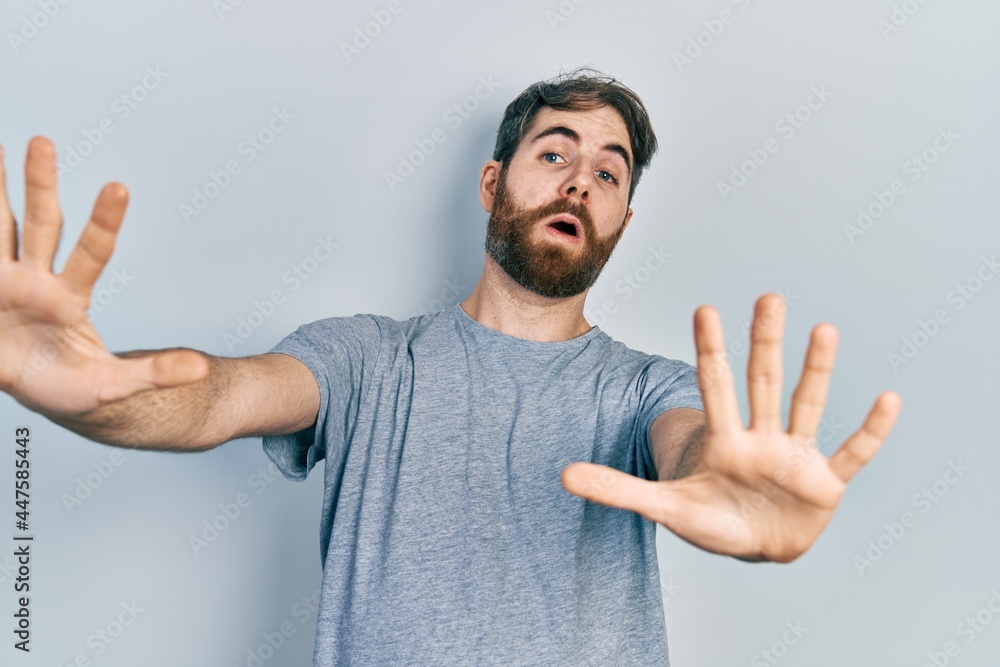 Caucasian man with beard wearing casual grey t shirt afraid and terrified with fear expression stop gesture with hands, shouting in shock. panic concept. Caucasian man with beard wearing casual grey t shirt afraid and terrified with fear expression stop gesture with hands, shouting in shock. panic concept.