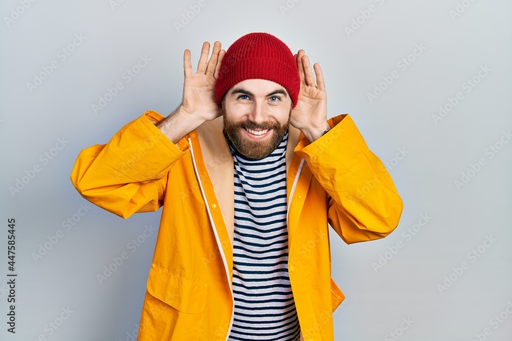 Caucasian man with beard wearing yellow raincoat smiling pulling ears with fingers, funny gesture. audition problem Caucasian man with beard wearing yellow raincoat smiling pulling ears with fingers, funny gesture. audition problem