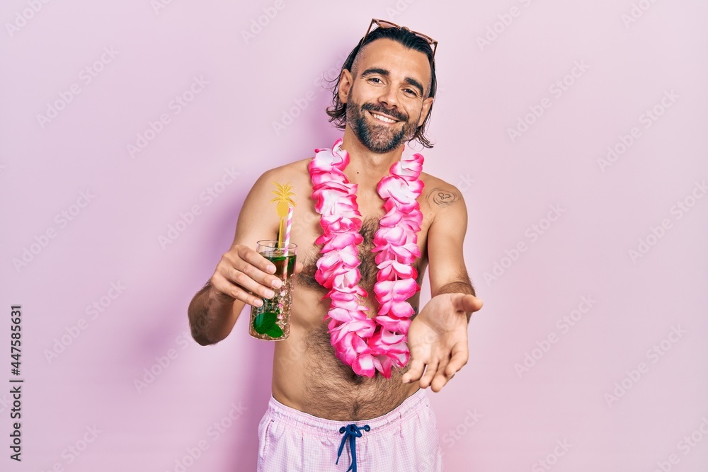 Young hispanic man wearing swimsuit and hawaiian lei drinking tropical cocktail smiling cheerful offering palm hand giving assistance and acceptance. Young hispanic man wearing swimsuit and hawaiian lei drinking tropical cocktail smiling cheerful offering palm hand giving assistance and acceptance.