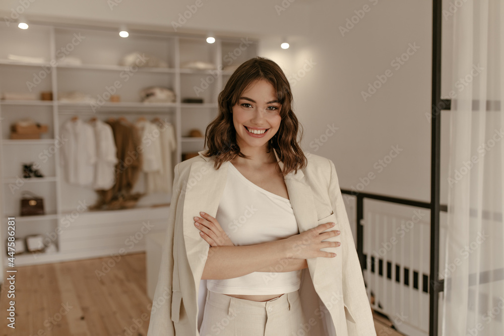 Cute brunette woman in linen pants, beige oversized jacket and white cropped top crosses arms and sincerely smiles in dressing room.