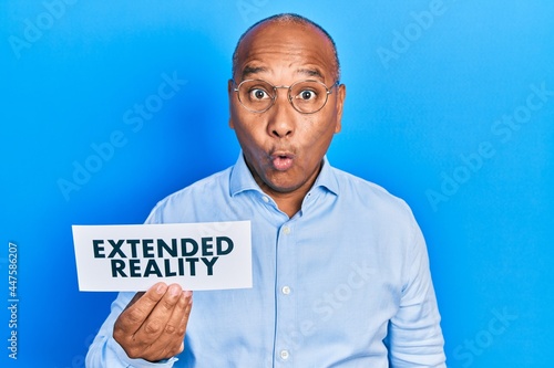 Middle age latin man holding paper with extended reality message scared and amazed with open mouth for surprise, disbelief face