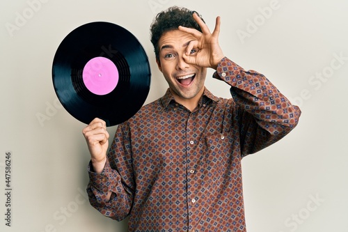 Young handsome man holding vinyl disc smiling happy doing ok sign with hand on eye looking through fingers